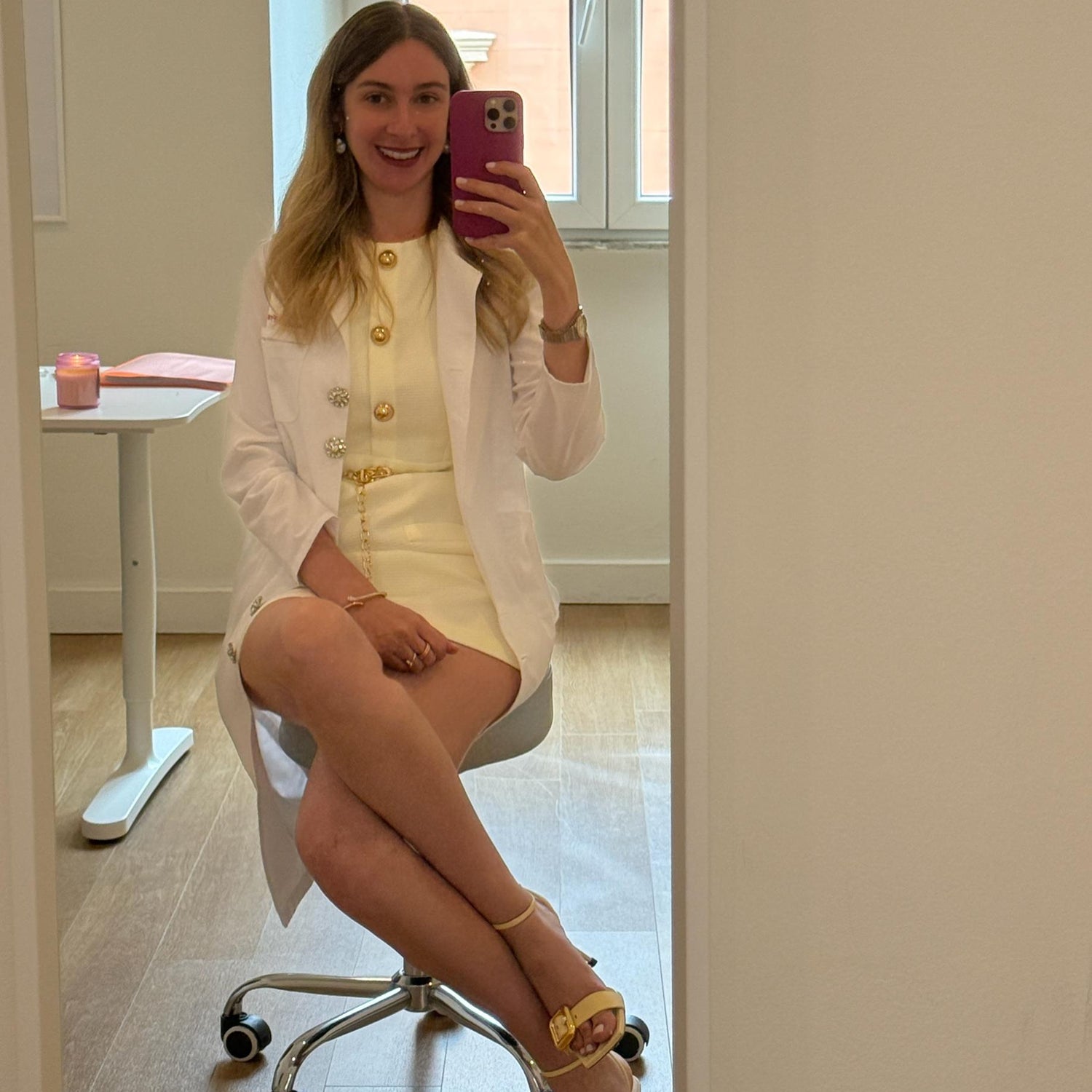 Woman taking a mirror selfie wearing a white blazer and yellow dress in a room with a window.